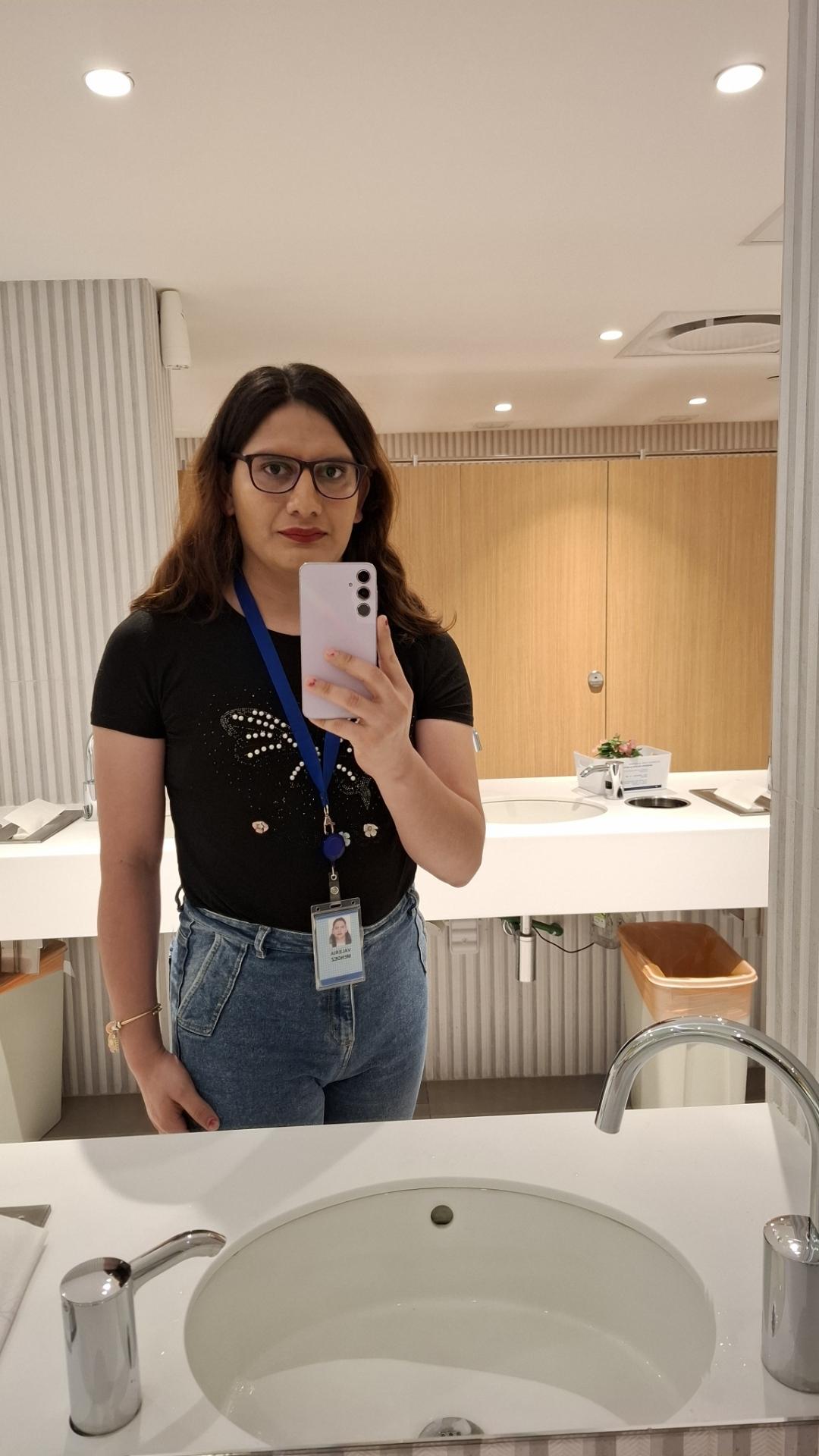 Foto de Val tomándose una selfie enfrente de un espejo. Cabello largo semiondulado de color castaño oscuro. Blusa negra de manga corta con un diseño de mariposa en pedrería de fantasía. Pantalón de mezclilla azul.